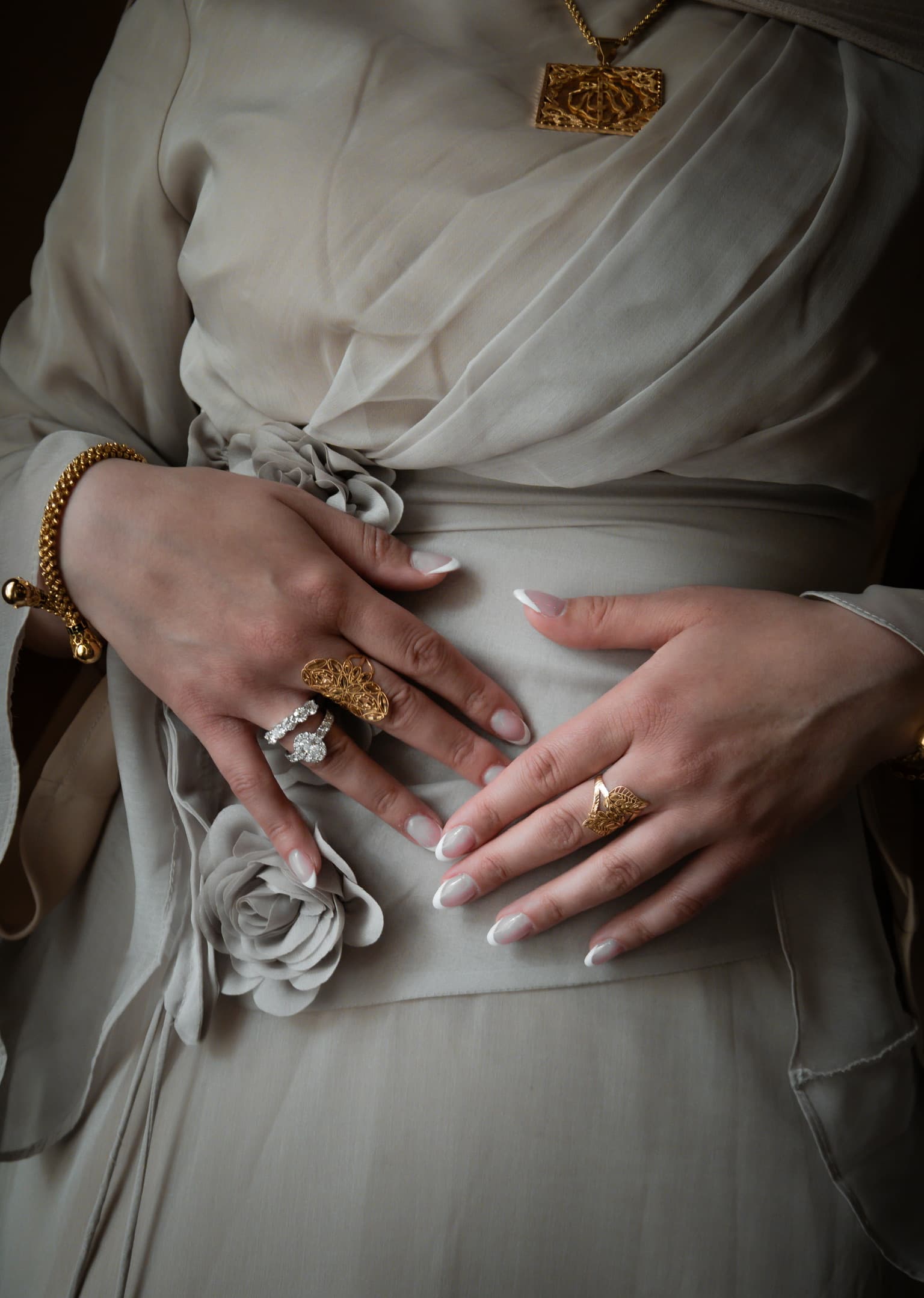 Close-up of clasped hands with engagement rings and a gold pendant necklace, photographed by ShuhraBilNoor