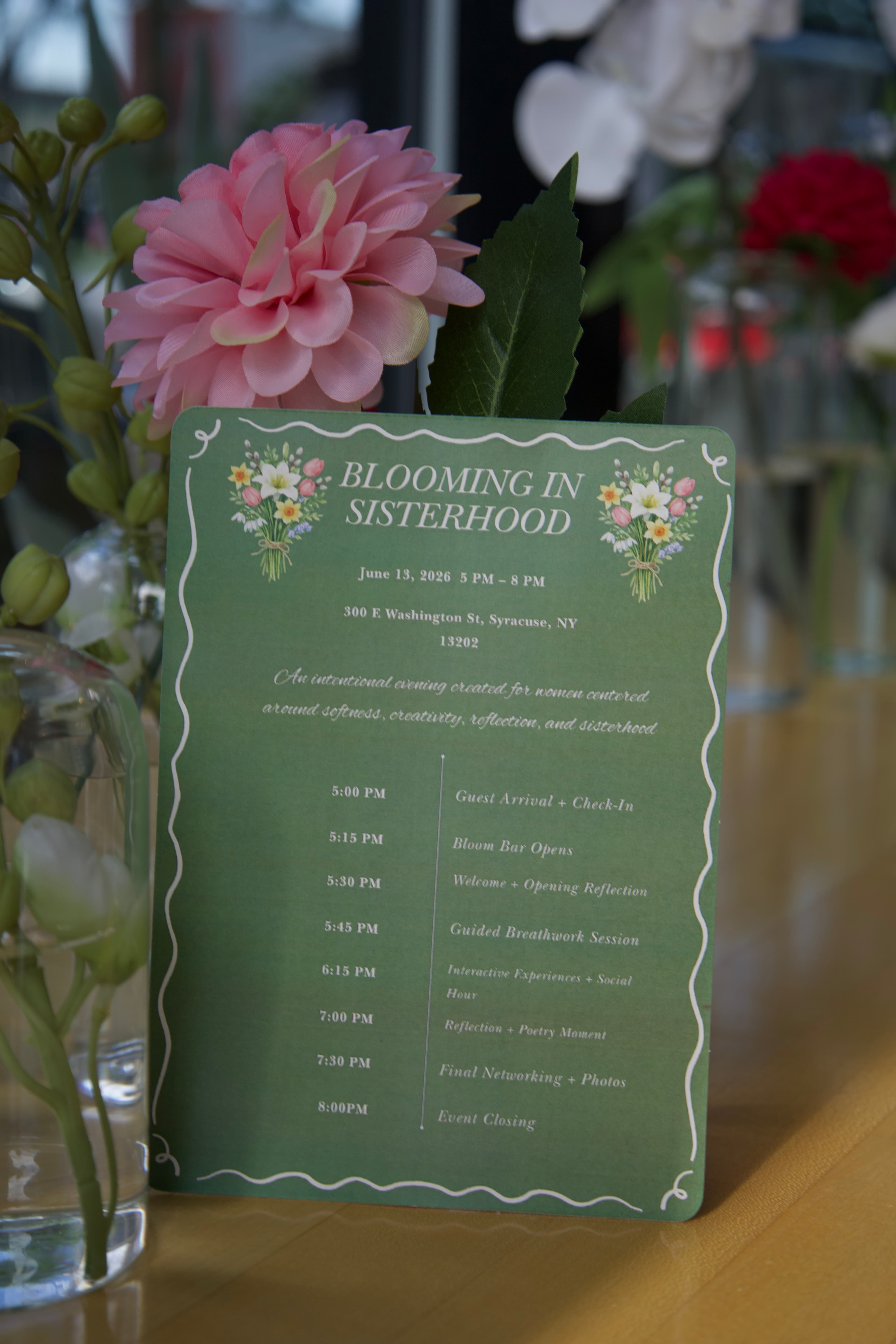 "Blooming in Sisterhood" event program card surrounded by flowers, photographed by ShuhraBilNoor