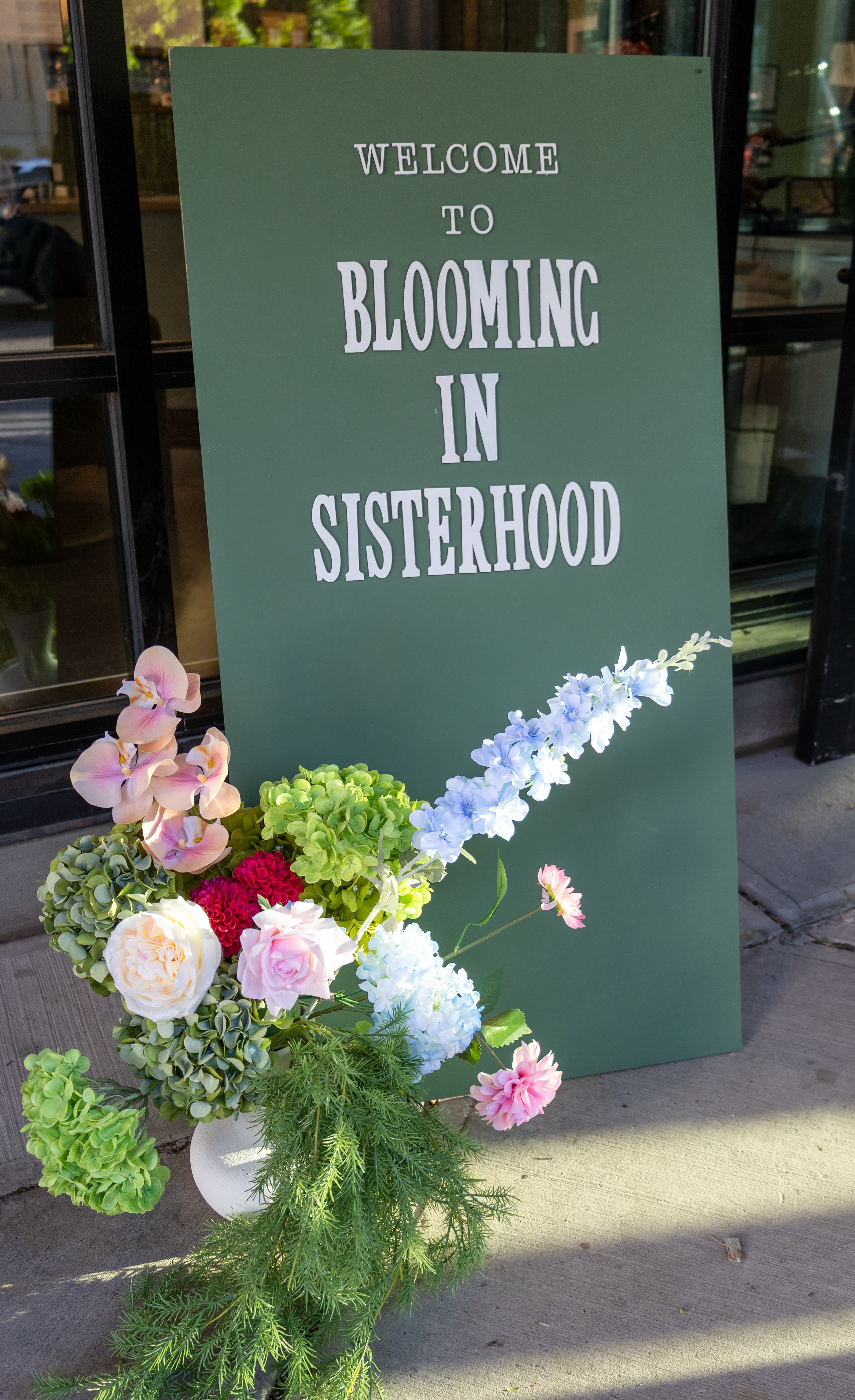 "Welcome to Blooming in Sisterhood" sign decorated with flowers outside the event venue, photographed by ShuhraBilNoor