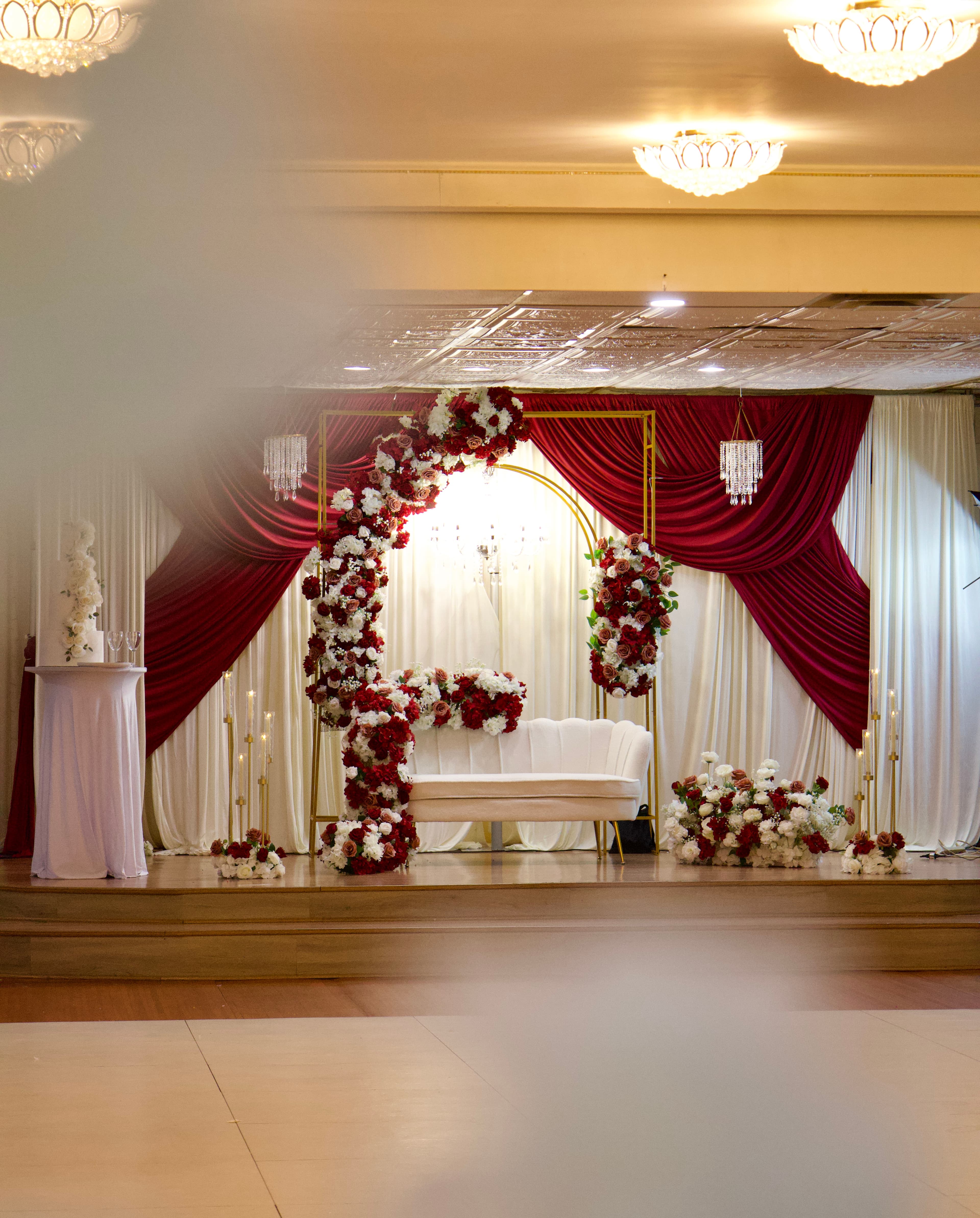 Reception stage decorated with red drapery, a floral arch, and a white loveseat, photographed by ShuhraBilNoor