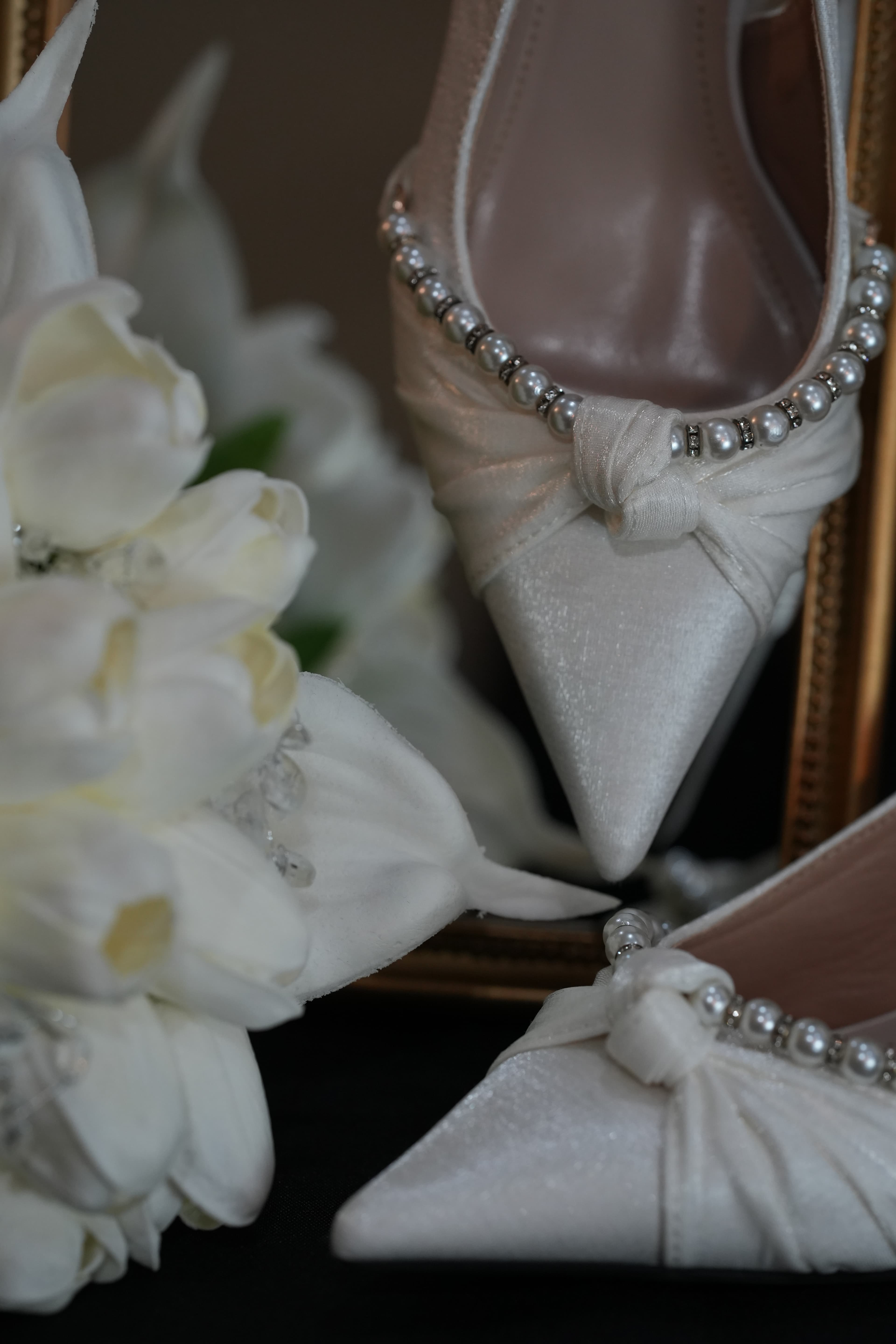 Close-up of pearl-embellished bridal shoes beside white floral bouquets, photographed by ShuhraBilNoor