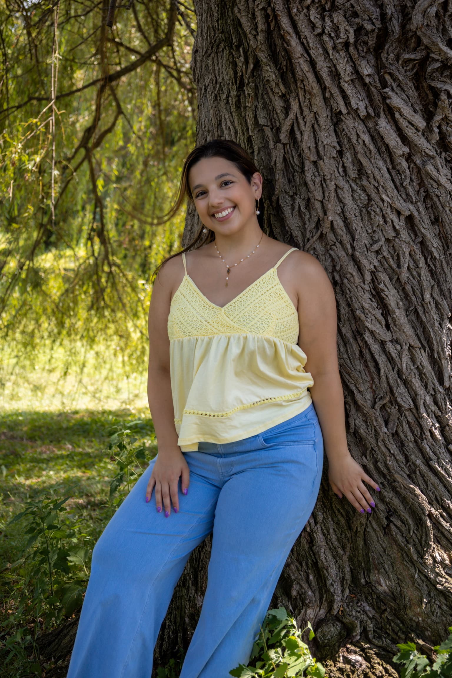 Full-length portrait photographed by ShuhraBilNoor, client smiling outdoors leaning against a tree