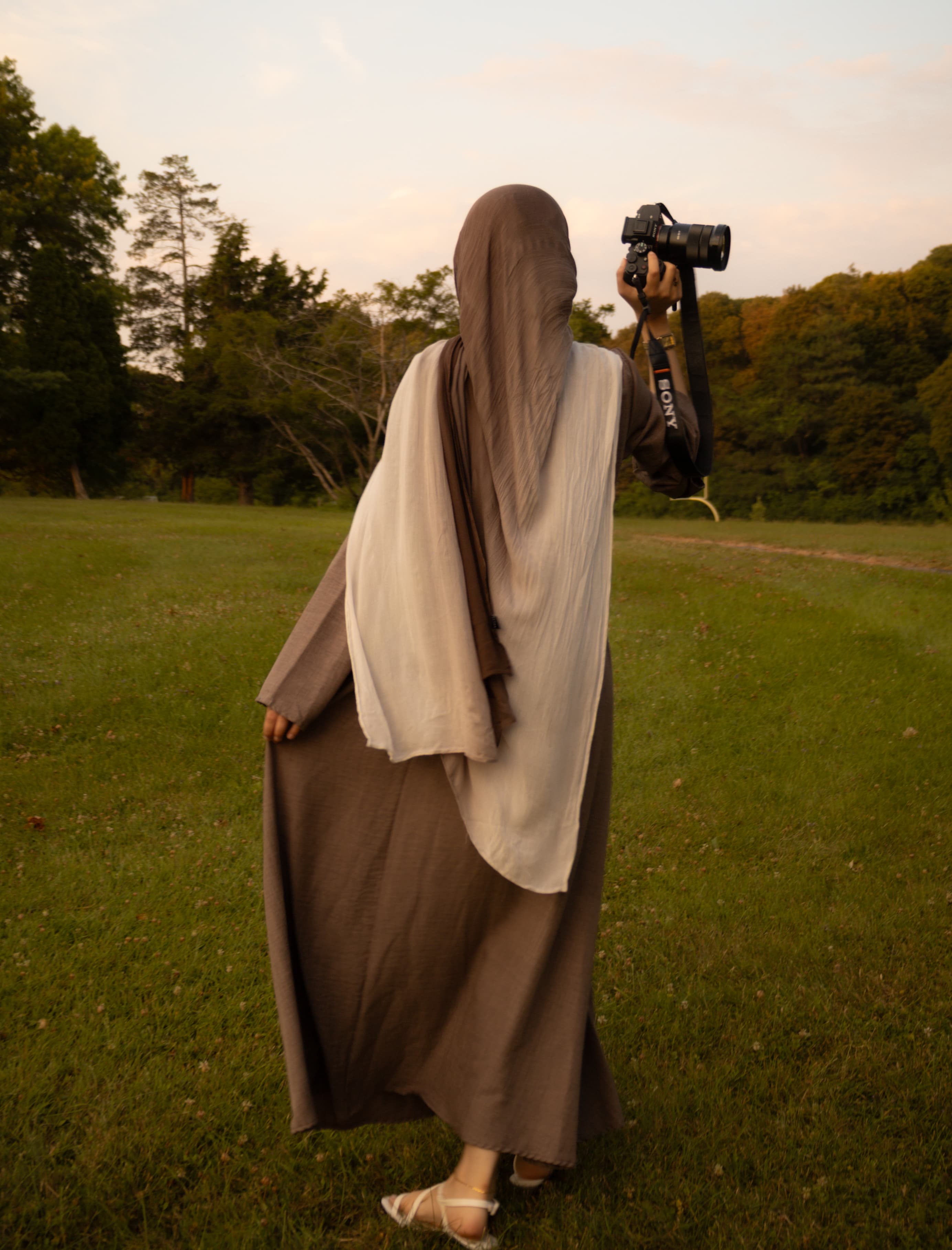Shahra, full-length, photographing outdoors in a grassy field, camera raised, seen from behind